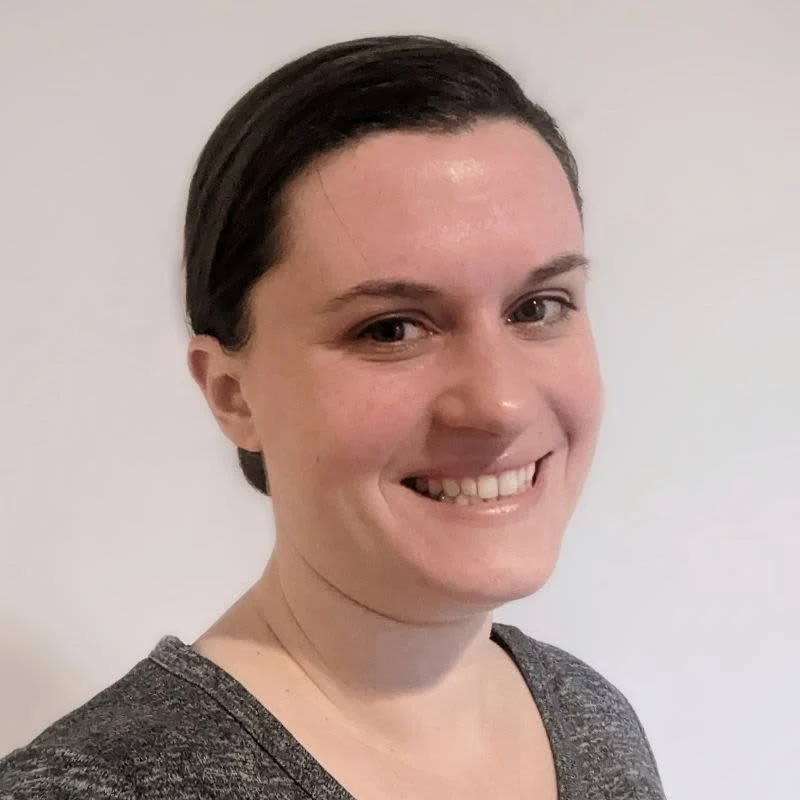 A woman in a gray shirt smiling in front of a white wall, raising awareness for mecfs.