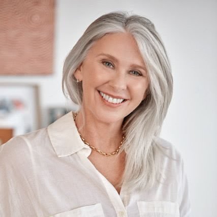 A woman with gray hair smiling in a white shirt and living with ME/CFS.